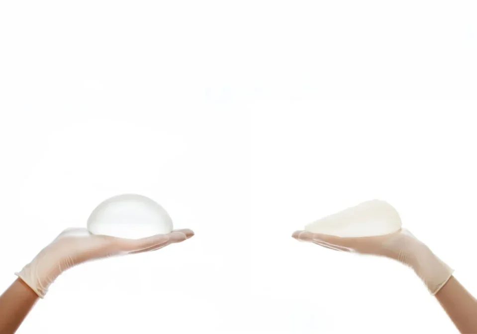 Two hands each holding a transparent silicone breast implant against a white background.